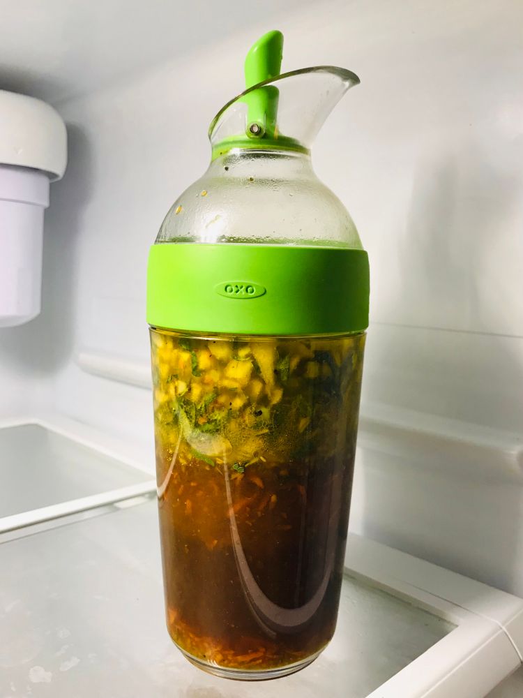 A transparent jar with a spout in a fridge. Inside it is a rich dressing with chunks of herbs and fruit.