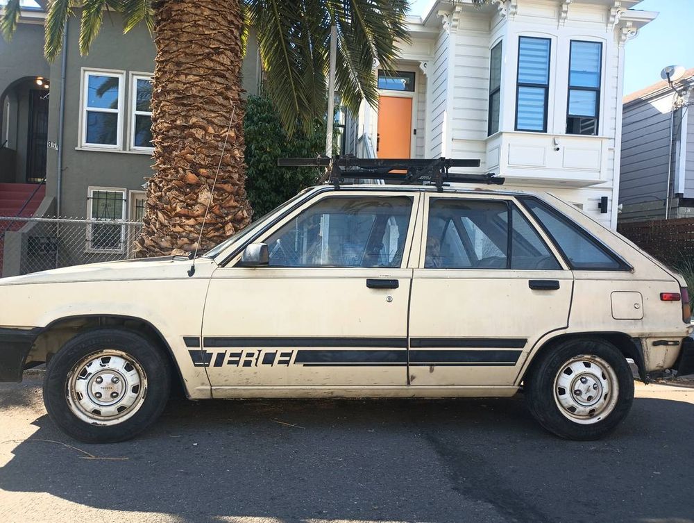 White 4-door 1984 hatchback Toyota Tercel, from the side, its hood pointed to the left of the frame and slightly cut off before the headlights. The Tercel occupies the entire horizontal length of the frame, its body dingy with faint mud splatters behind the front wheel. The rims are shiny white and newer than the rest of the car. It has a bike stand on top. In the background, there are 2 nice-looking townhomes (dark green, and white with bright yellow door), a chainlink fence and the trunk of a palmetto tree positioned in front of the dark green house on the left (behind windshield/hood of car).