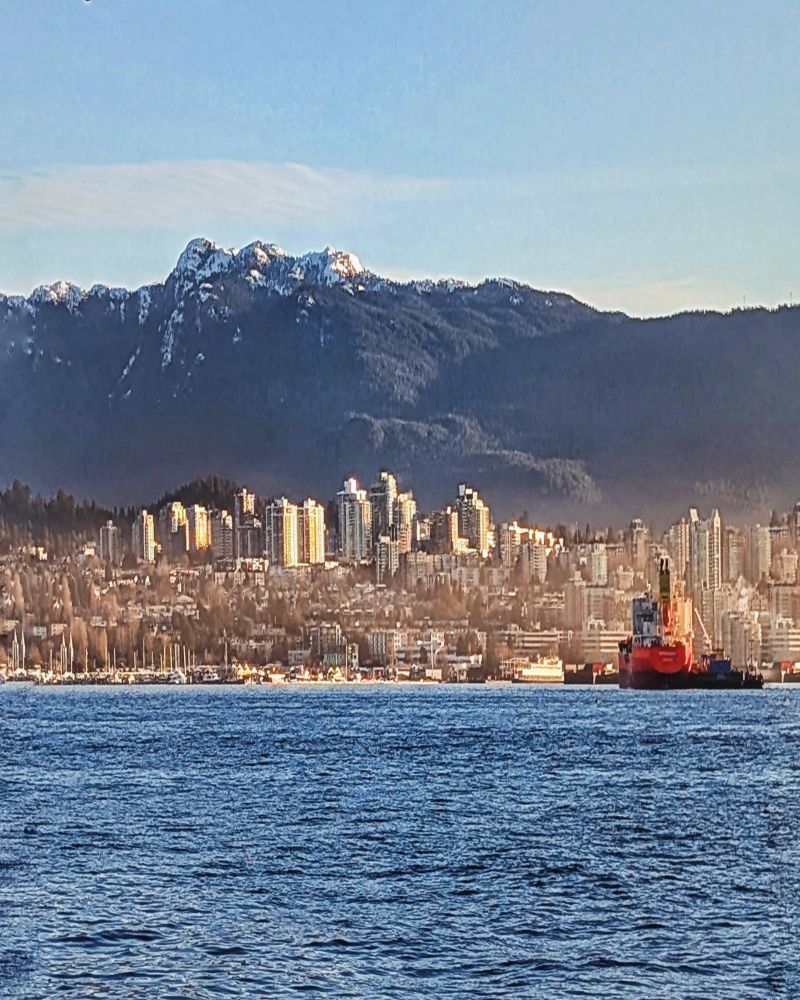A mountain is behind downtown North Vancouver buildings. Water in the foreground is the Vancouver Harbour.