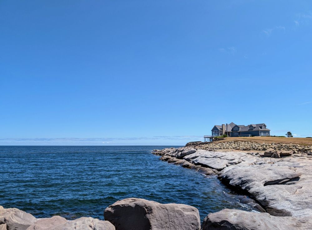 A rocky shore looking out over the Atlantic ocean. A large home sits just above the horizon.