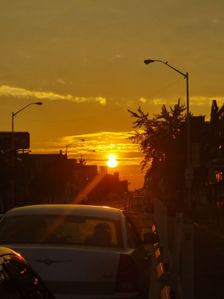 Shot from a city street, the image shows the rising sun creating a lens flare over buildings and the car in the foreground. Street lights help frame the photo 