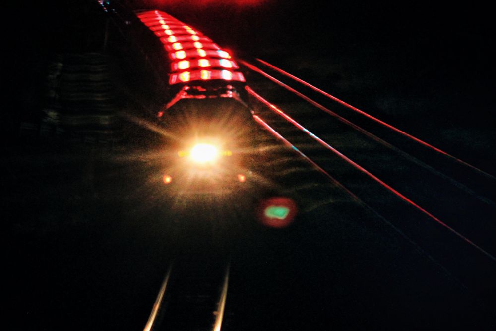 A night shot of a train, its top and the tracks lit in red from a large signal light stand it has just passed under. 