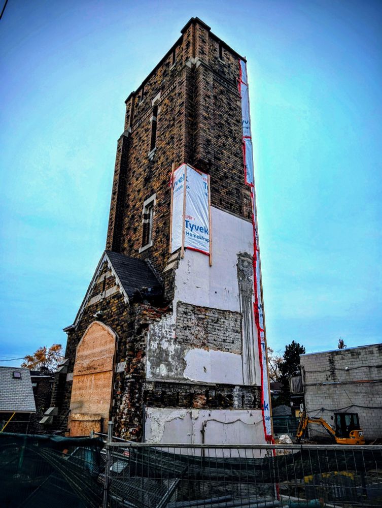 A church undergoing extensive renovations has only the front entrance and steeple remaining. The front door is covered with plywood.