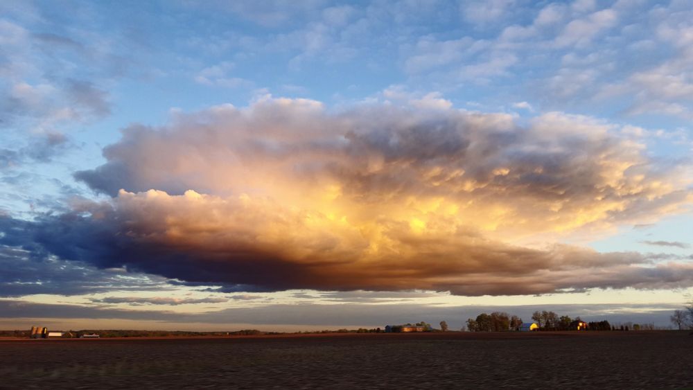 A massive cloud dwarfing a farm is laden with moisture while its center gets lit up from the setting sun.