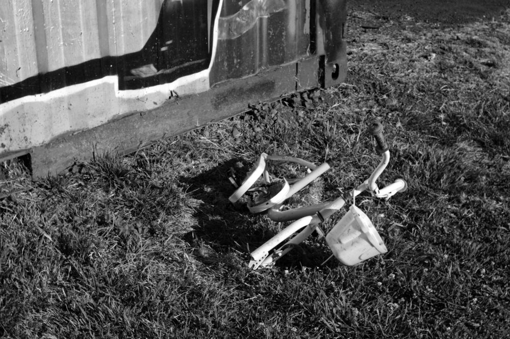 A black and white photo of a child's bicycle, with a basket, stripped of most of it's parts and laying in grass next to a graffiti covered shipping container.
