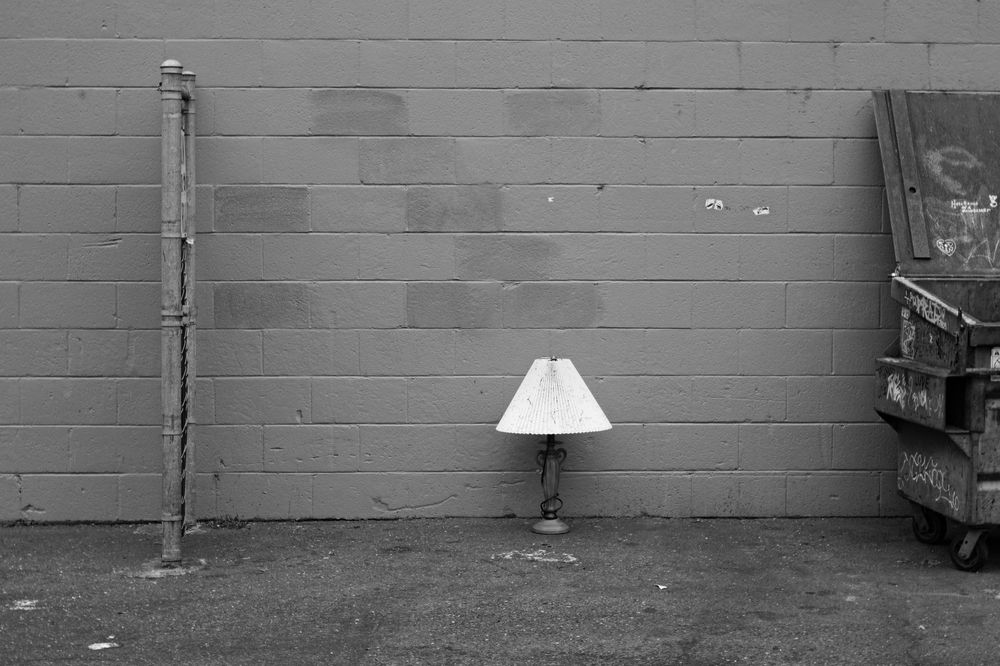 A black and white photo of a lamp standing on the asphalt of a parking lot, with a brick wall behind it, between a section of fence and a graffiti covered dumpster.