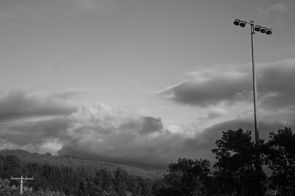 A black and white photo of clouds seen above distant hills with trees in between, with some power and light poles in view.