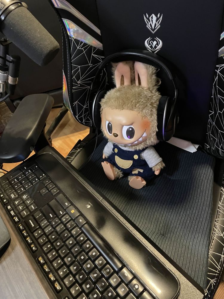 Time To Chill labubu is a larger doll. She sits in the middle of a K/DA Secret Labs gaming chair atop a black seat cushion with large black headphones on both sides of her head (it is worth noting she has rabbit-like ears on top of her head). In front of her is a computer keyboard and to the top left corner of the screen an SM7B Shure microphone is visible