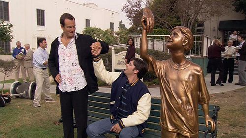 GOB Bluth and Steve Holt from Arrested Development clasping hands while standing next to a bronze statue of a kid holding a hold with no one attached to the hand