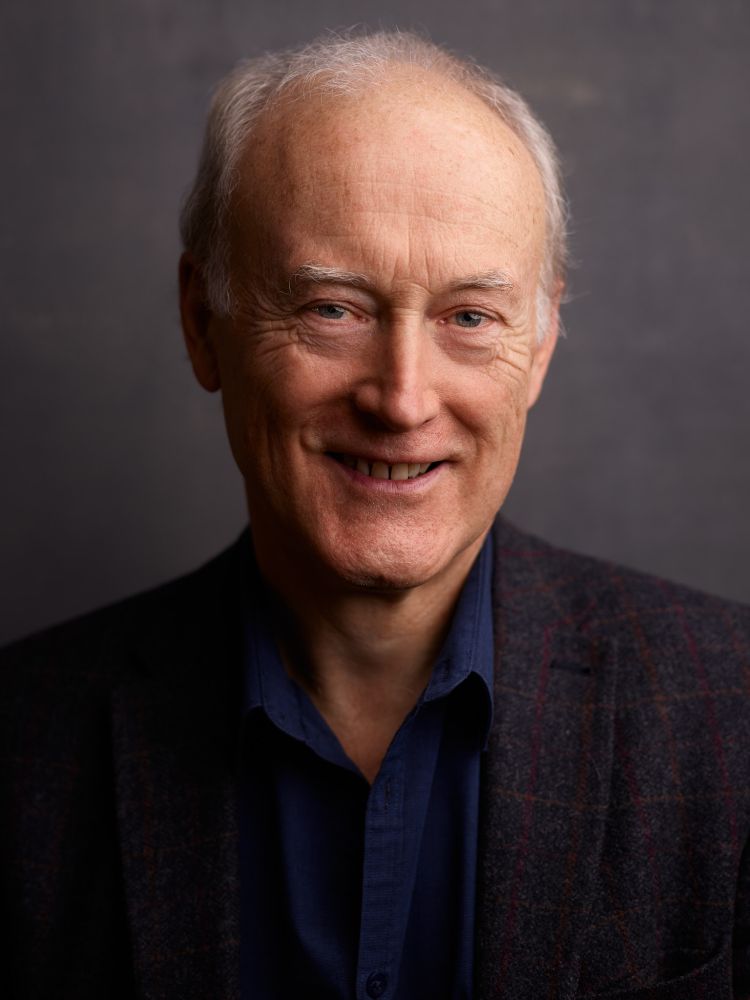 A head and shoulders shot of Nicholas Graham. He is looking into the camera and smiling. Nick is wearing a black suit jacket and dark blue shirt. He has white hair and light brown eyes. Behind him is a dark grey background.