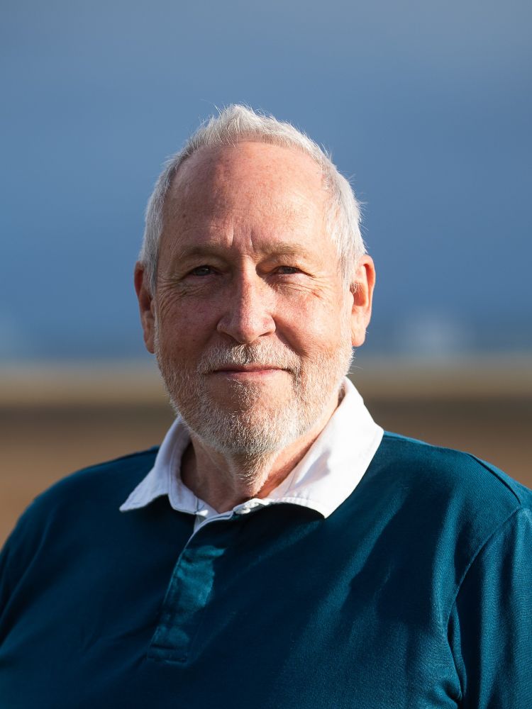 A man with neat white hair and a white beard smiles at the camera. He is wearing a petrol blue rugby style shirt with a white collar. He is smiling gently at the camera. His head and shoulders are visible and behind him the blurred background is varying shades of sand brown and sky blue.