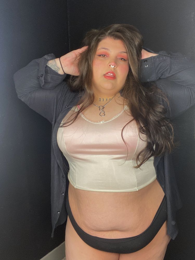 Curvy plus size woman stand playing with her hair looking at the camera while in a long unbuttoned black shirt and a dainty silk top. 
