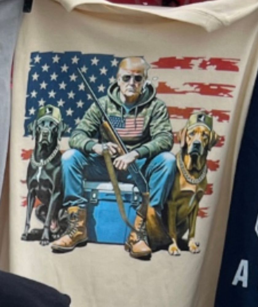 A shirt depicting trump in an olive drab hoodie with an American flag on it. He's holding a pump action shotgun, sitting on a cooler, and flanked by two dogs which are wearing baseball caps and gold chains. He's wearing jeans and work boots and he's in front of an American flag. 