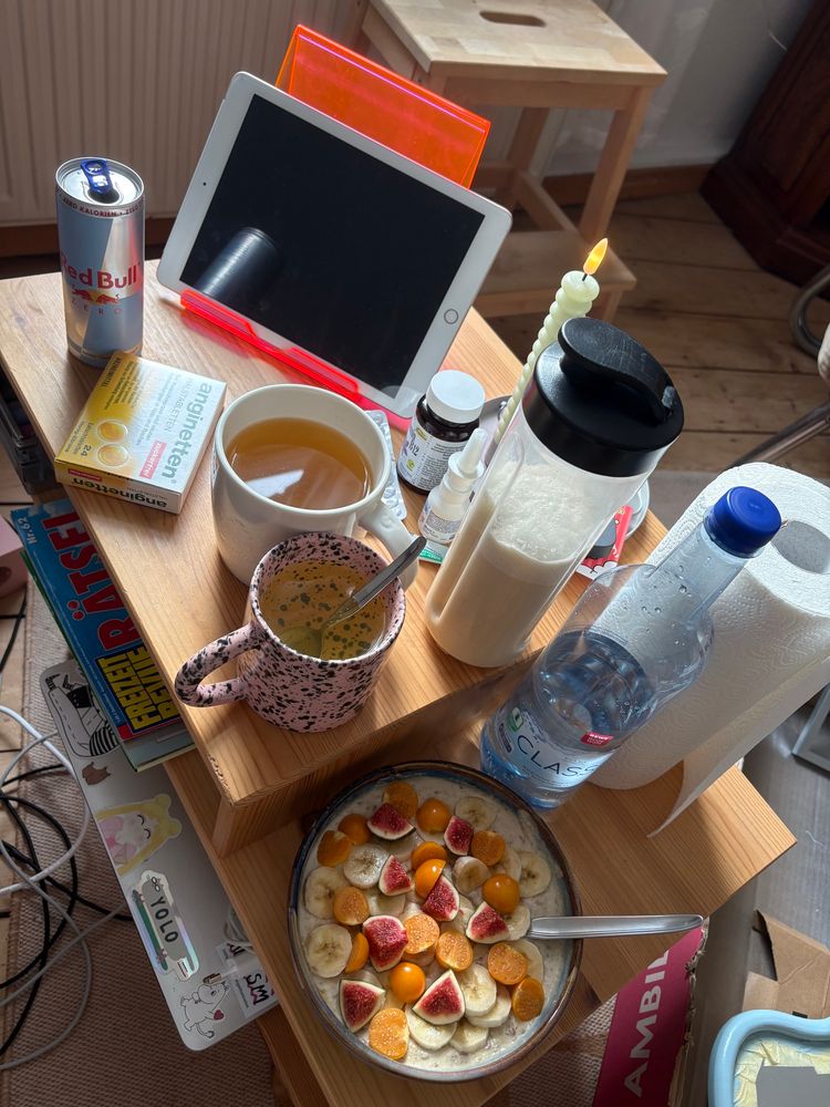 auf einem kleinen Couchtisch stehen 2 Tassen, eine Flasche Wasser, ein Milchshake, eine Dose RedBull, verschiedene Medikamente, eine Schüssel Porridge mit Obst, eine Rolle Küchenpapier, eine Kerze und ein iPad; im Hintergrund Chaos 
