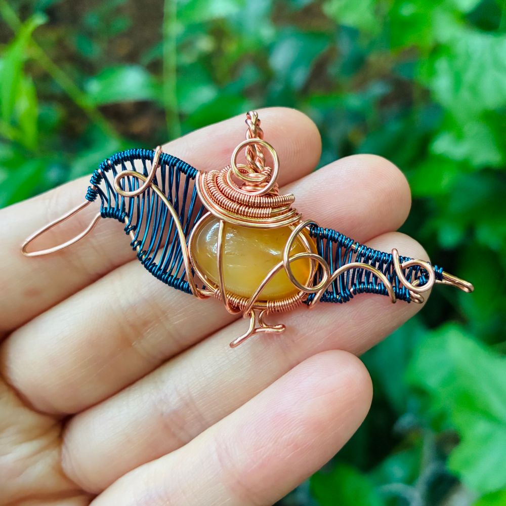 Copper wirewrapped pendant in the shape of a kingfisher with an orange stone body, blue weaved head/tail, and a tiny wire foot