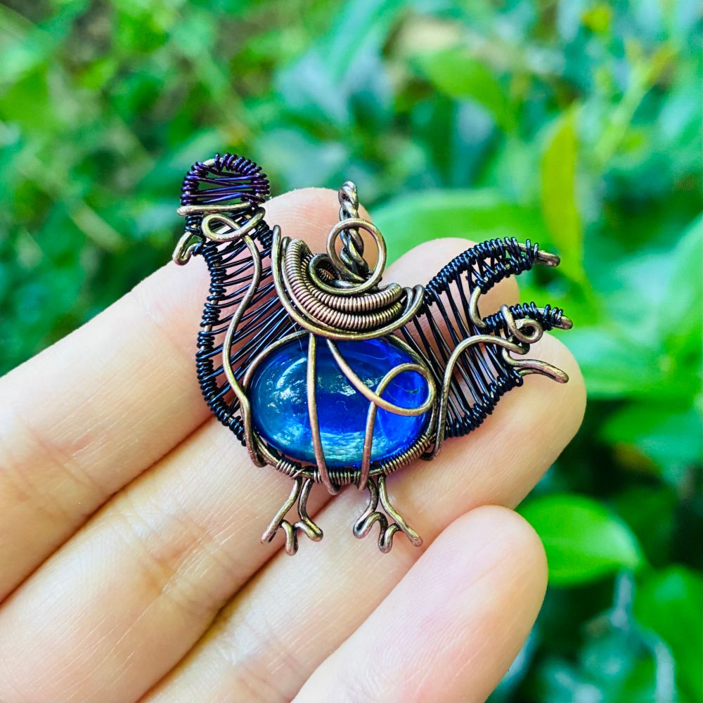 copper wirewrapped pendant in the shape of a chicken with a flashy blue stone body, dark blue weaved head/tail, purple comb, and 2 tiny wire feet