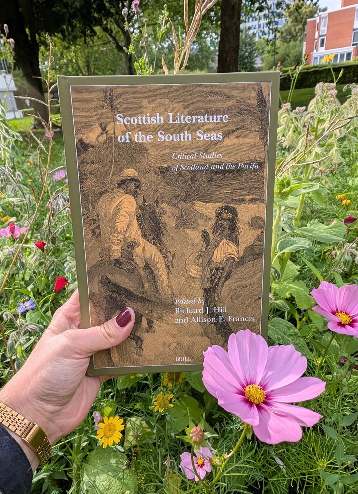 Holding the book "Scottisch Literature of the South Seas" amidst a meadow in bloom, with a brightly pink flower in the foreground