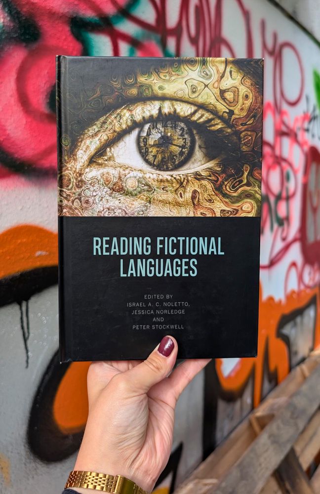Holding the essay collection "Reading Fictional Languages", edited by Noletto, Norledge & Stockwell, in front of a wall covered in hardly legible graffiti writing in orange and red
