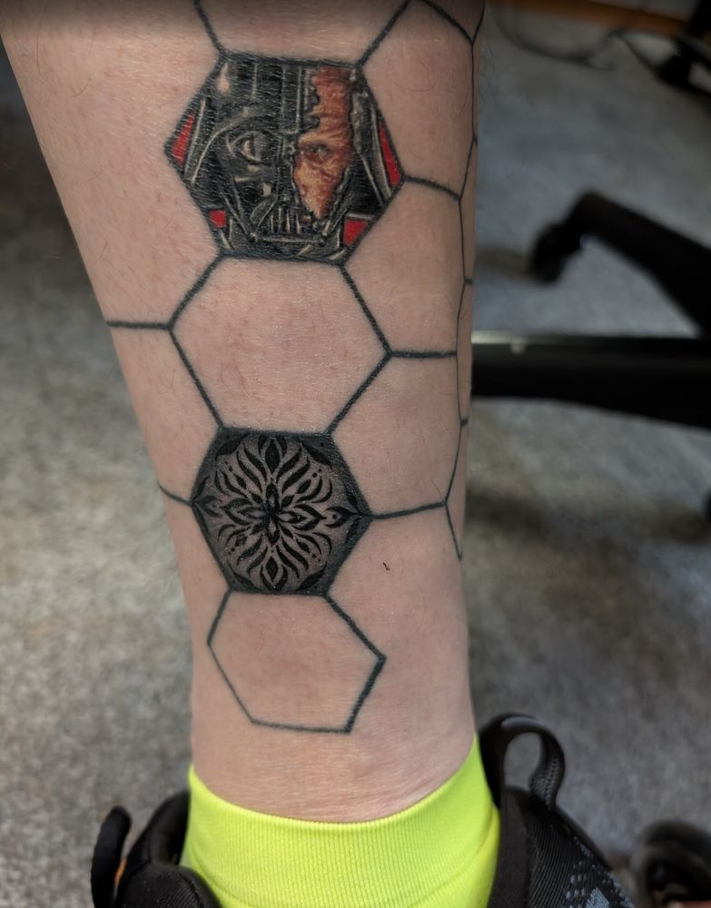 Side photo of lower leg. The leg has some connected hexagons.
Within one of these hexagons is a tattoo of Darth Vader. Half of his face is covered by his helmet, the other half is open and shows his disfigured (burned) face from Return of the Jedi.
Another hexagon has a set of geometric shapes in black and grey - as if someone did tie dye but with just black dye.