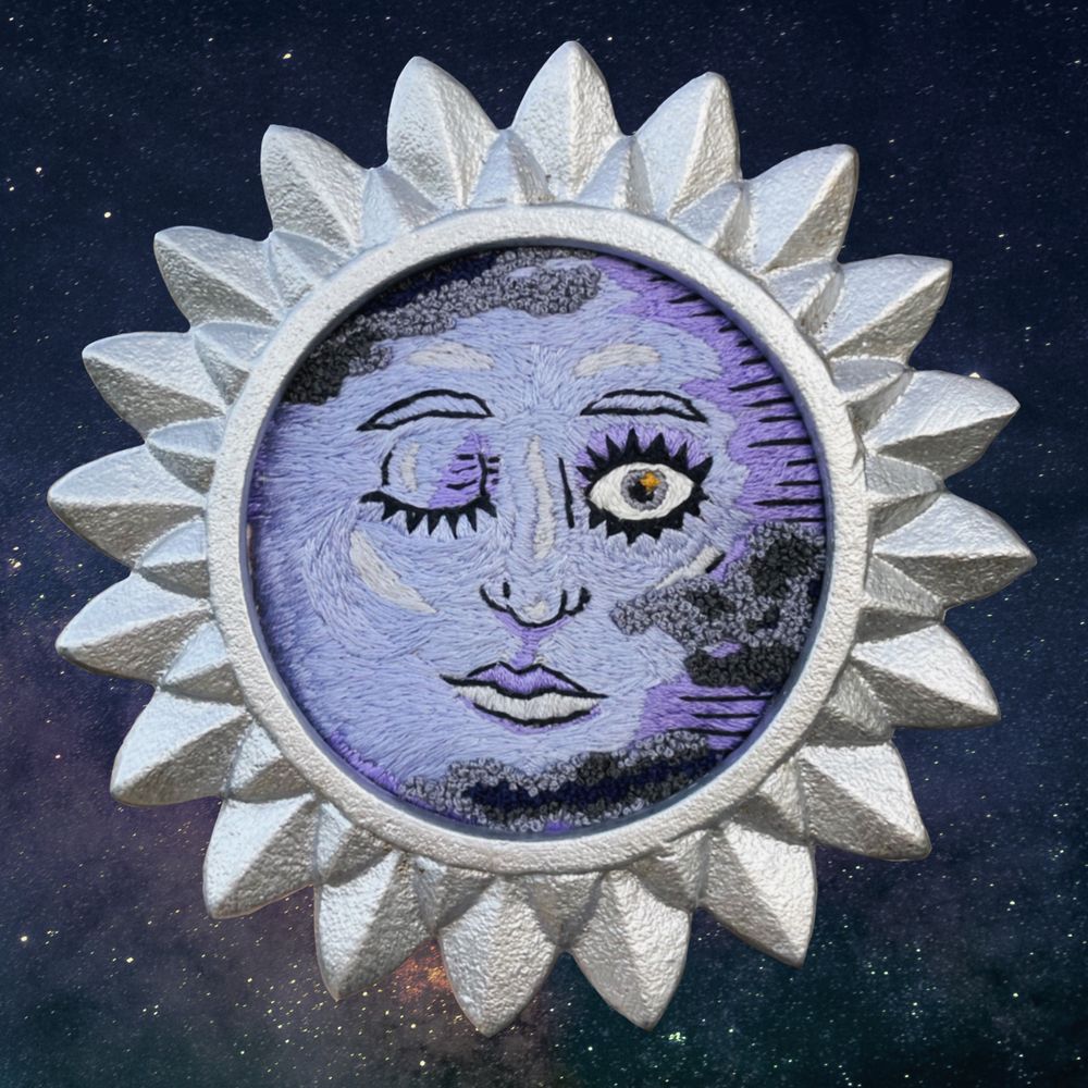 An embroidered face within a silver sun frame to look like the moon. The right eye is open and the left eye is closed. There are clouds embroidered on the top left and bottom right of the face. 