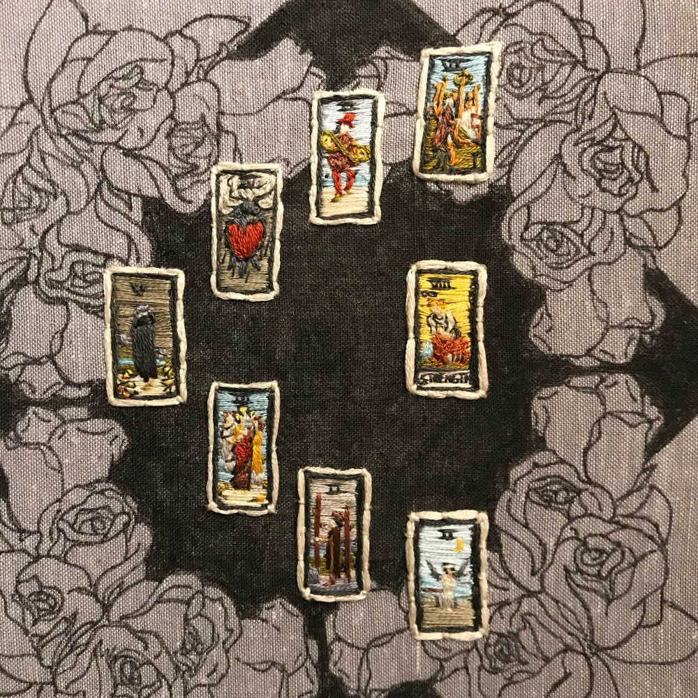 Eight embroidered tiny tarot cards in a half moon spread. Going from the top counter clockwise is the 6 of wands, 2 of pentacles, 3 of swords, 5 of cups, 3 of cups, 2 of wands and 2 of swords. The inner card is strength.
