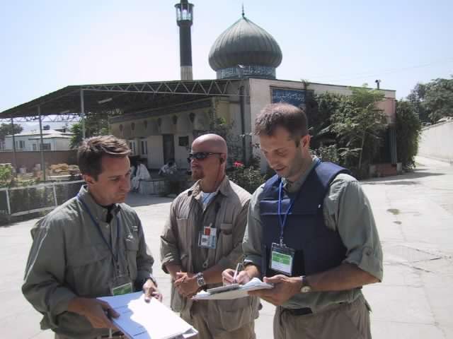 Three individuals in professional attire holding papers and engaging in a discussion outside a building with a minaret and a dome visible in the background.