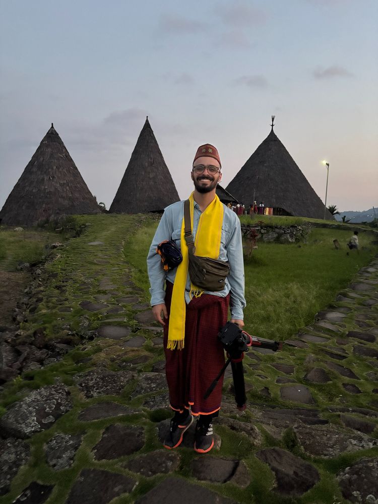 Person wearing a cap, glasses, and traditional sarong standing on a stone path at dusk, with the unique conical structures of the Todo village in the background.