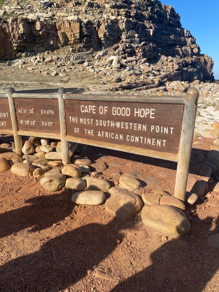 Wooden signs at the Cape of Good Hope, labeled as the most south-western point of the African continent, with rocky terrain and a mountain in the background.