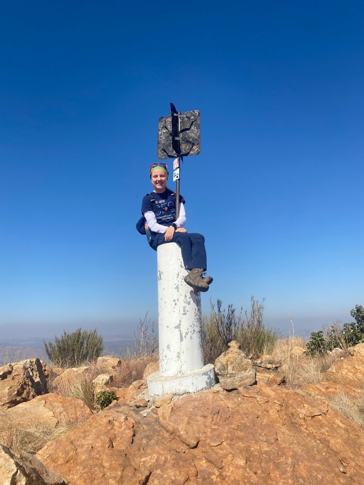 Person sitting on a summit marker at the top of a mountain, with a clear blue sky in the background.