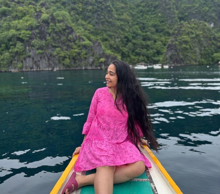 Person in a pink dress sitting at the front of a boat, overlooking the serene waters and lush green cliffs of Manila.