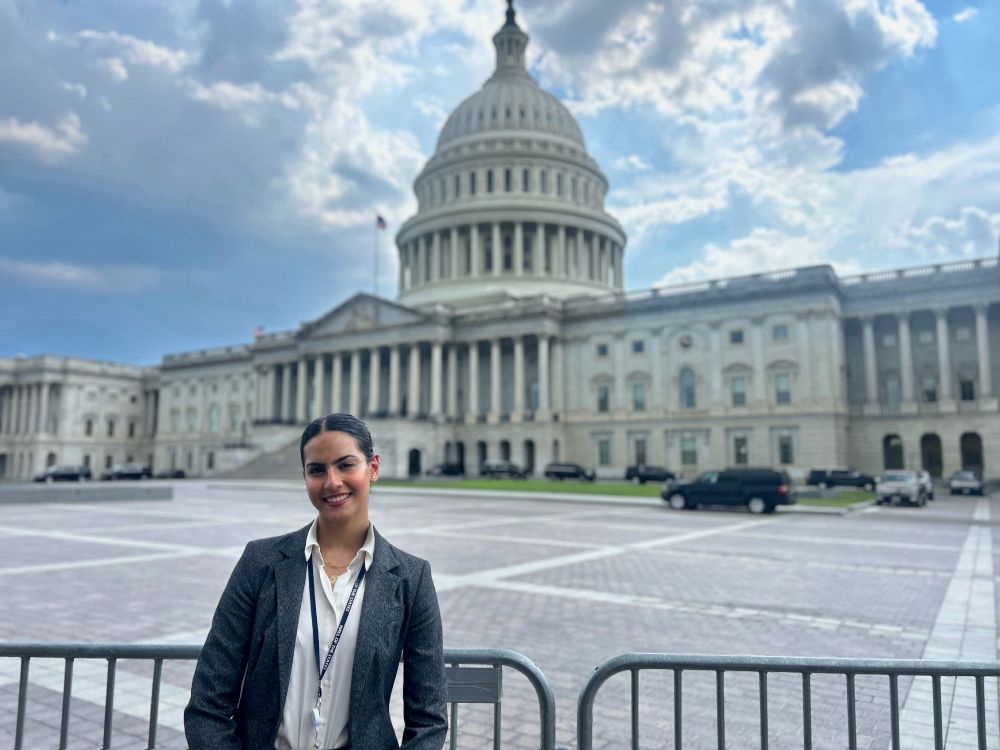 A person wearing a business suit stands in front of the United States Capitol building under a partly cloudy sky.