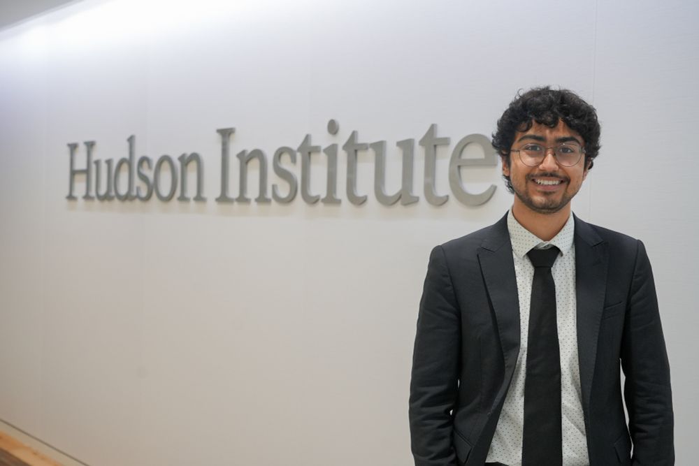 A person with curly hair and glasses, wearing a suit, smiles in front of a sign reading "Hudson Institute".