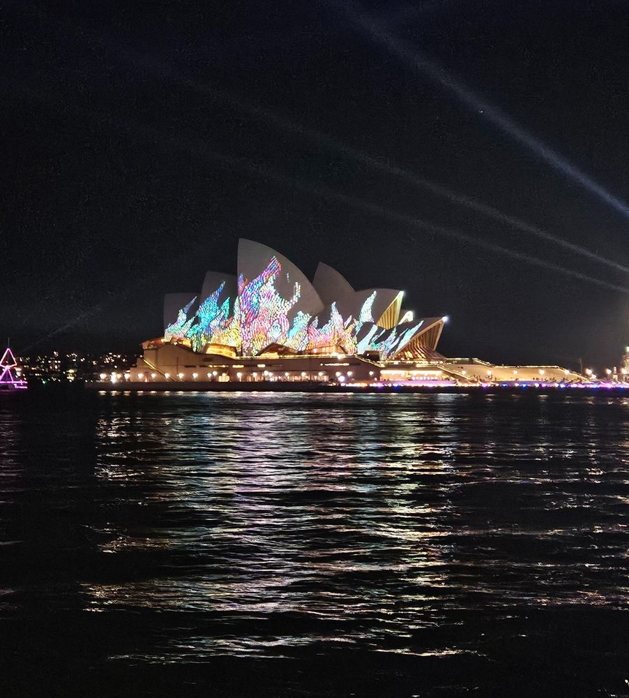 Night view of the Sydney Opera House illuminated with colorful light projections, reflected in the water below.