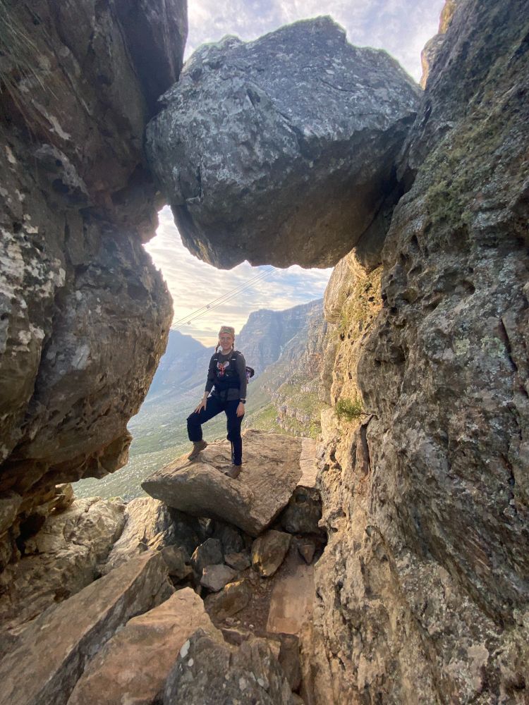 A person standing between large boulders with a scenic mountain backdrop.