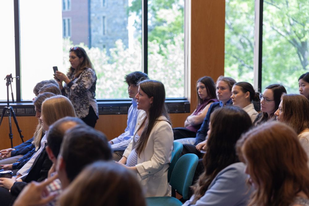 Attendees listening to a speaker at a professional seminar, with some participants checking their phones, in a conference room with large windows.