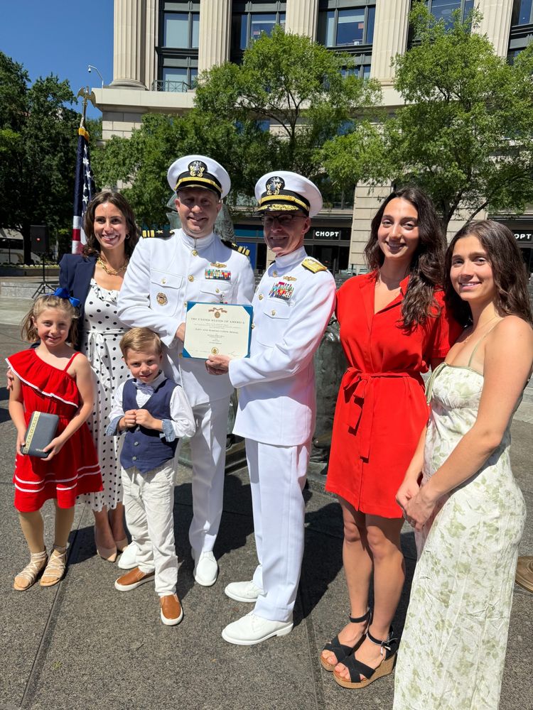 Group of seven people celebrating an award ceremony for a naval officer in uniform, with three adults and four children standing together, smiling in front of a building on a sunny day.
