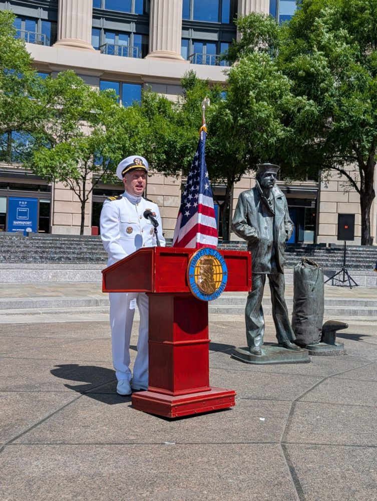 A person in a white naval uniform stands at a podium with the seal of the United States, giving a speech next to a U.S. flag and a statue of a standing figure in casual attire.