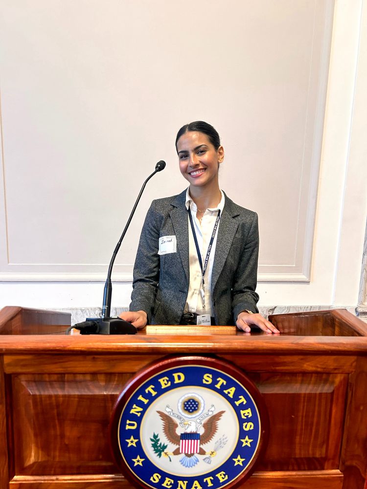 A person stands at a podium with the United States Senate seal, smiling and wearing a business suit with a name badge.
