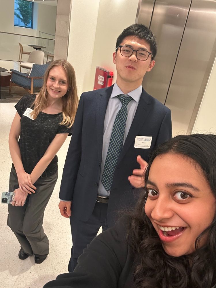 Three professionals posing for a selfie in a lobby, with one person in the foreground and the other two slightly behind, all wearing business attire.