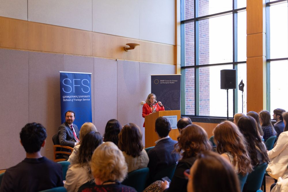 A person is giving a speech at a podium with the Georgetown University School of Foreign Service logo visible, in a room with seated attendees and large windows.