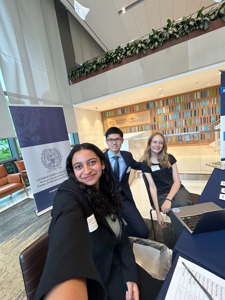 Three individuals smiling at a registration table inside a building, with a banner reading "Georgetown University" in the background.