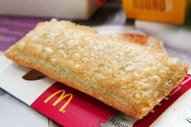 Das Bild zeigt ein frittiertes Apfeltaschen-Dessert von McDonald's. Es liegt auf seiner Verpackung, die rot-weiß ist und das gelbe McDonald's-Logo trägt. Die Apfeltasche hat eine goldbraune, knusprige Oberfläche mit kleinen Blasen, die vom Frittieren stammen. Im Hintergrund sind unscharf andere Verpackungen und ein Tablett zu erkennen.