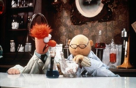 Das Bild zeigt Beaker und Dr. Bunsen Honeydew, zwei bekannte Figuren aus der Muppet-Show, die in einem Labor stehen. Links ist Beaker zu sehen, eine hochgewachsene, orangefarbene Puppe mit roten Haaren, großem Nasenknopf und weit aufgerissenen Augen. Er trägt einen weißen Laborkittel und eine schwarze Krawatte. Rechts daneben steht Dr. Bunsen Honeydew, eine kleinere, rundliche Puppe mit hellbeiger Haut, Glatze und runder Brille. Auch er trägt einen weißen Laborkittel und ist gerade damit beschäftigt, mit einem Mörser und Stößel weiße Substanzen zu zermahlen. Auf dem Labortisch stehen mehrere Glasgeräte, darunter ein Kolben mit blauer Flüssigkeit. Im Hintergrund sind Regale mit Gläsern und Flaschen zu sehen, sowie eine altmodische Tapete und ein gerahmtes Porträt an der Wand. Die Szene vermittelt den Eindruck eines humorvollen, etwas altmodischen Chemielabors.