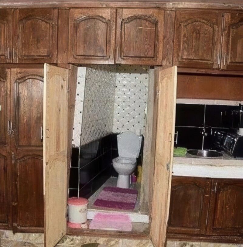 In einer Küchenzeile aus dunklem Holzschränken wurde eine Schranktür geöffnet, hinter der sich überraschenderweise eine Toilette verbirgt. Der kleine Raum ist gefliest und enthält eine weiße Toilette, eine rosa Badematte, einen passenden kleinen Teppich sowie einen Abfalleimer. Alles ist windschief und sieht gebastelt aus.