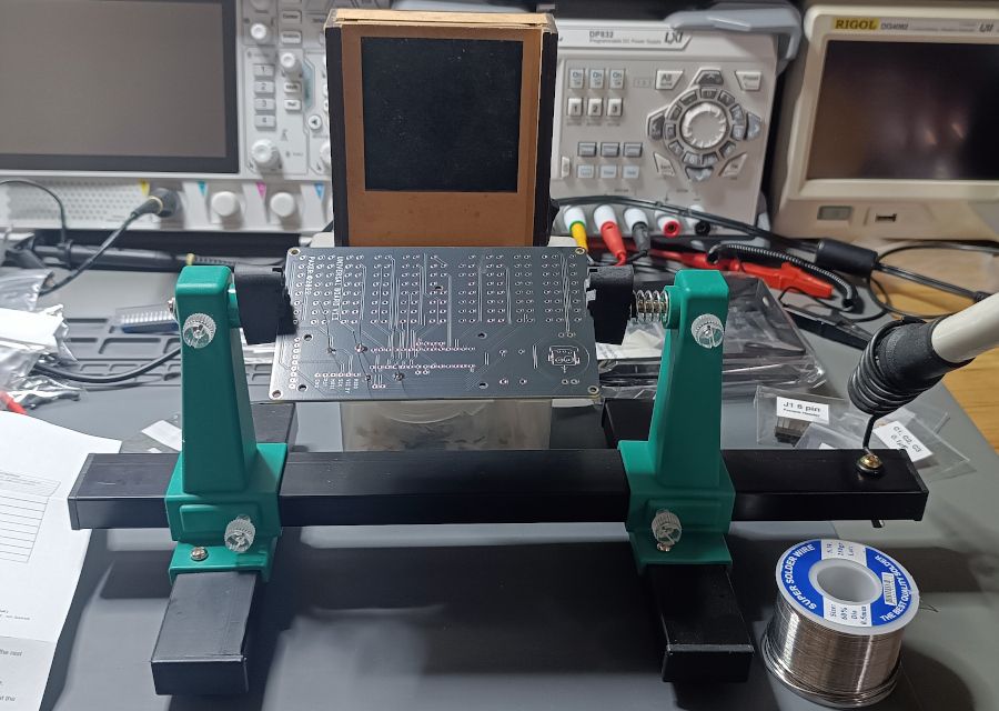 The bare PCB mounted on a soldering vise with a couple of resistors soldered already. There's a hand made fume extractor at the back and the bags with the components and the instructions can be seen around it.