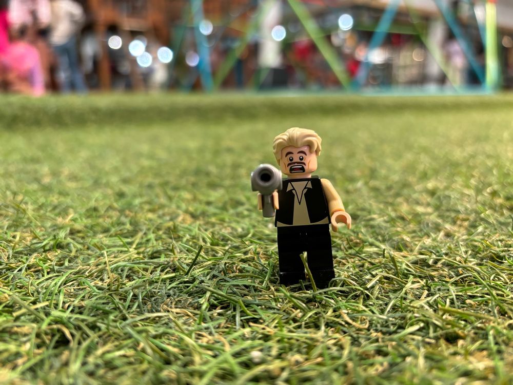 Lego mini figure standing in grass with surprise look on face, blonde hair, white shirt black vest, black pants and holding a sci-fi blaster. 