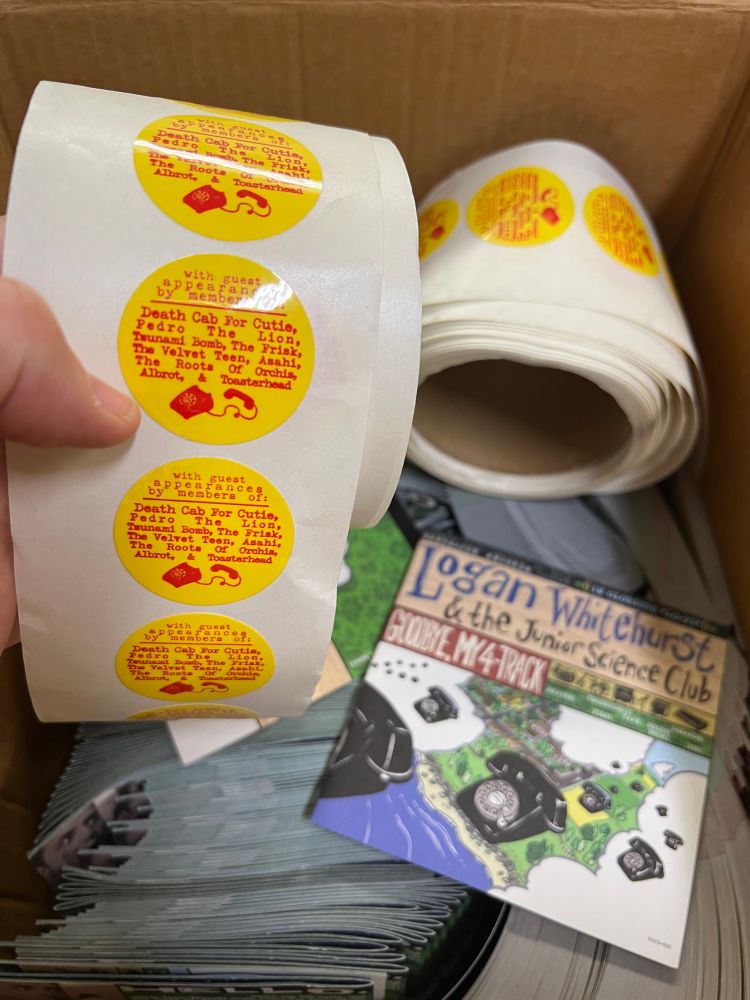 Hand holding a roll of round yellow stickers with red text. Hype stickers from the Logan Whitehurst & The Junior Science Club Goodbye, My 4-Track CD original pressing. 
Text reads: 
“With guest appearances by members of: Death Cab For Cutie, Pedro The Lion, Tsunami Bomb, The Frisk, The Velvet Teen, Asahi, The Roots Of Orchis, Albrot, & Toasterhead.”