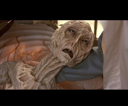 A picture of an alien prop from a movie or TV show, showing an anthropomorphic aliens face and upper shoulders staring up at someone else imploringly while reclining on said person's arm.

The alien is dessicated and leathery, with a single tooth and large yellow eyes.