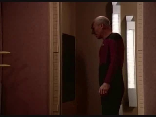 A screenshot of captain Picard from star trek (a bald white man in a red and black bodysuit uniform) standing in a warmly lit red residential setting with futuristic feel. He is staring at a black alcove in the wall, a replicator from the show.