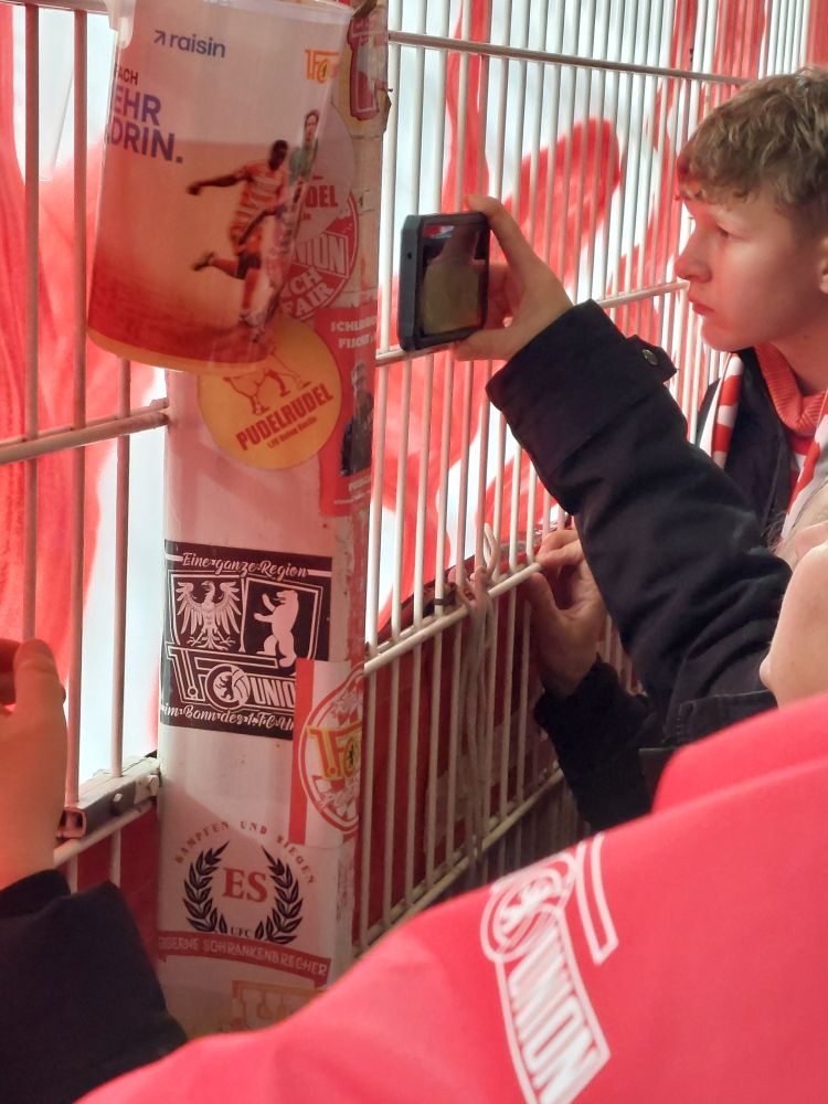 Im Stadion ist die Sicht durch ein Banner verhängt, Leute gucken auf dem Handy das Spiel.